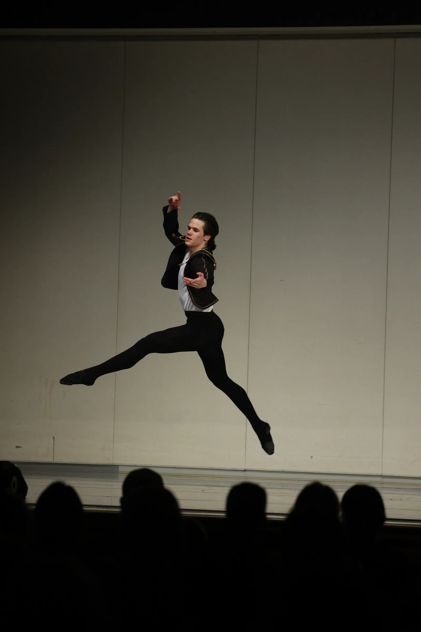A male ballet dancer in a black jacket and tights soars mid-leap, legs fully split, against a pale backdrop above silhouetted audience heads.