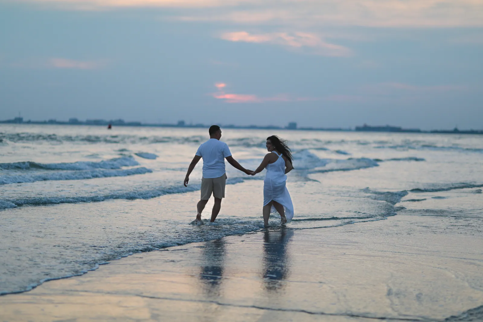 A couple holding hands wades into shallow surf at dusk, she in a flowing white dress, with pink-tinged clouds and a distant shoreline beyond.