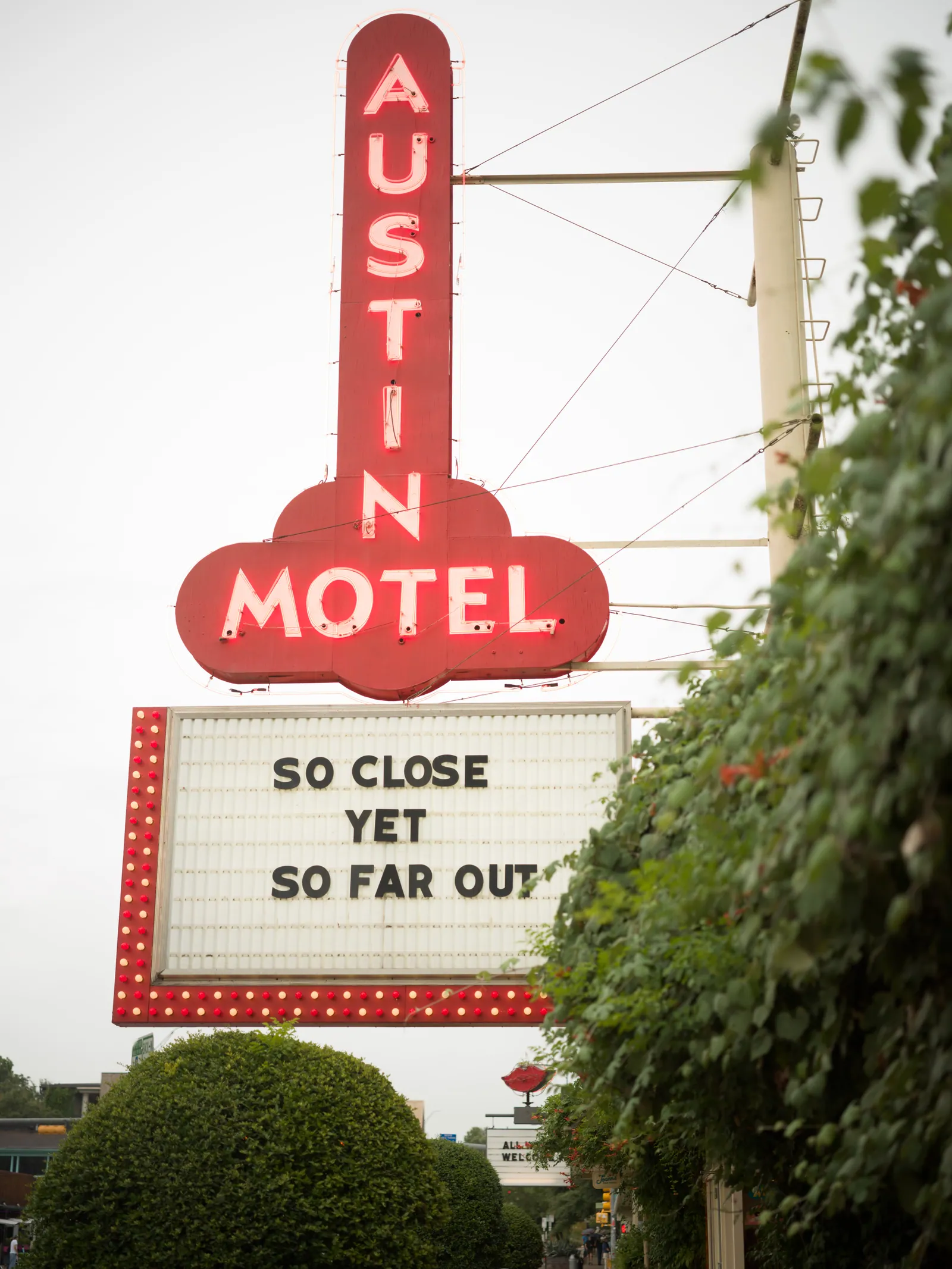 A red neon Austin Motel sign rises above a marquee reading So Close Yet So Far Out, framed by green foliage against an overcast sky.