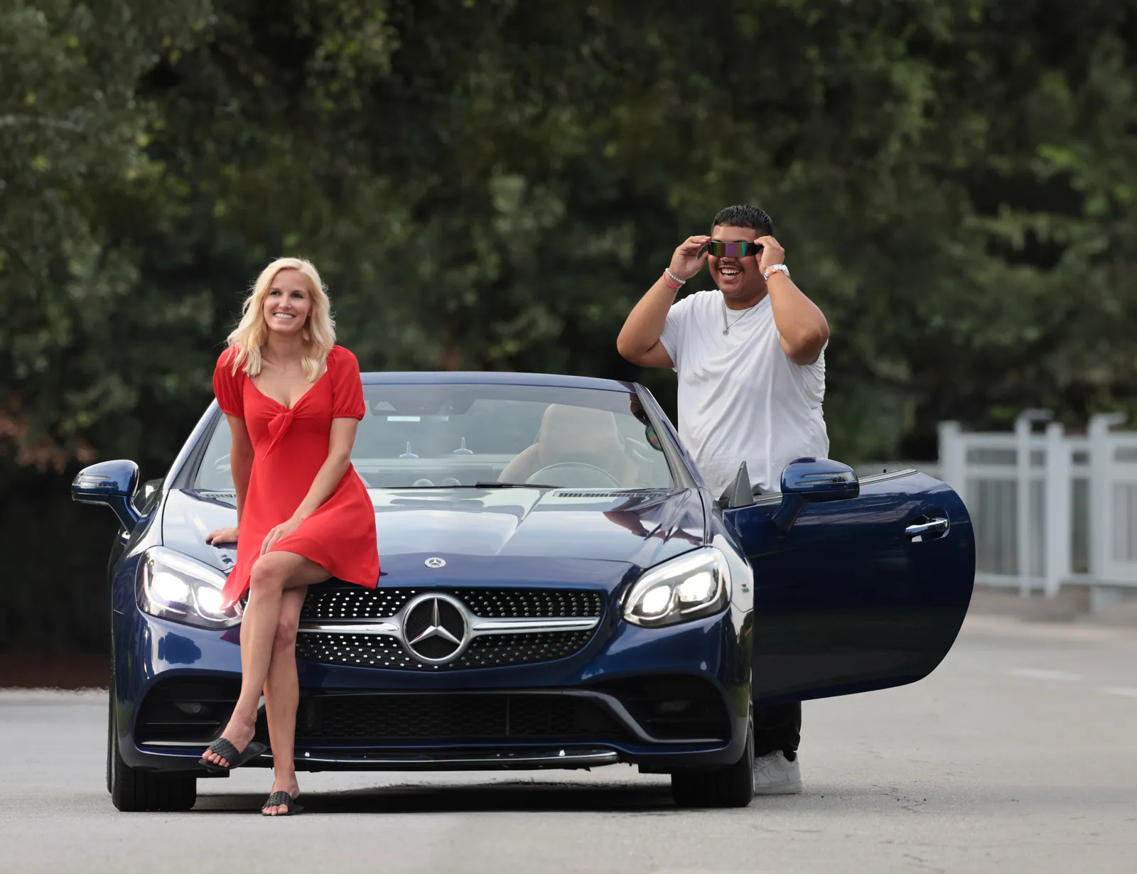 A woman in a red dress perches on the hood of a dark blue Mercedes convertible while a man in white adjusts reflective sunglasses by the open door.