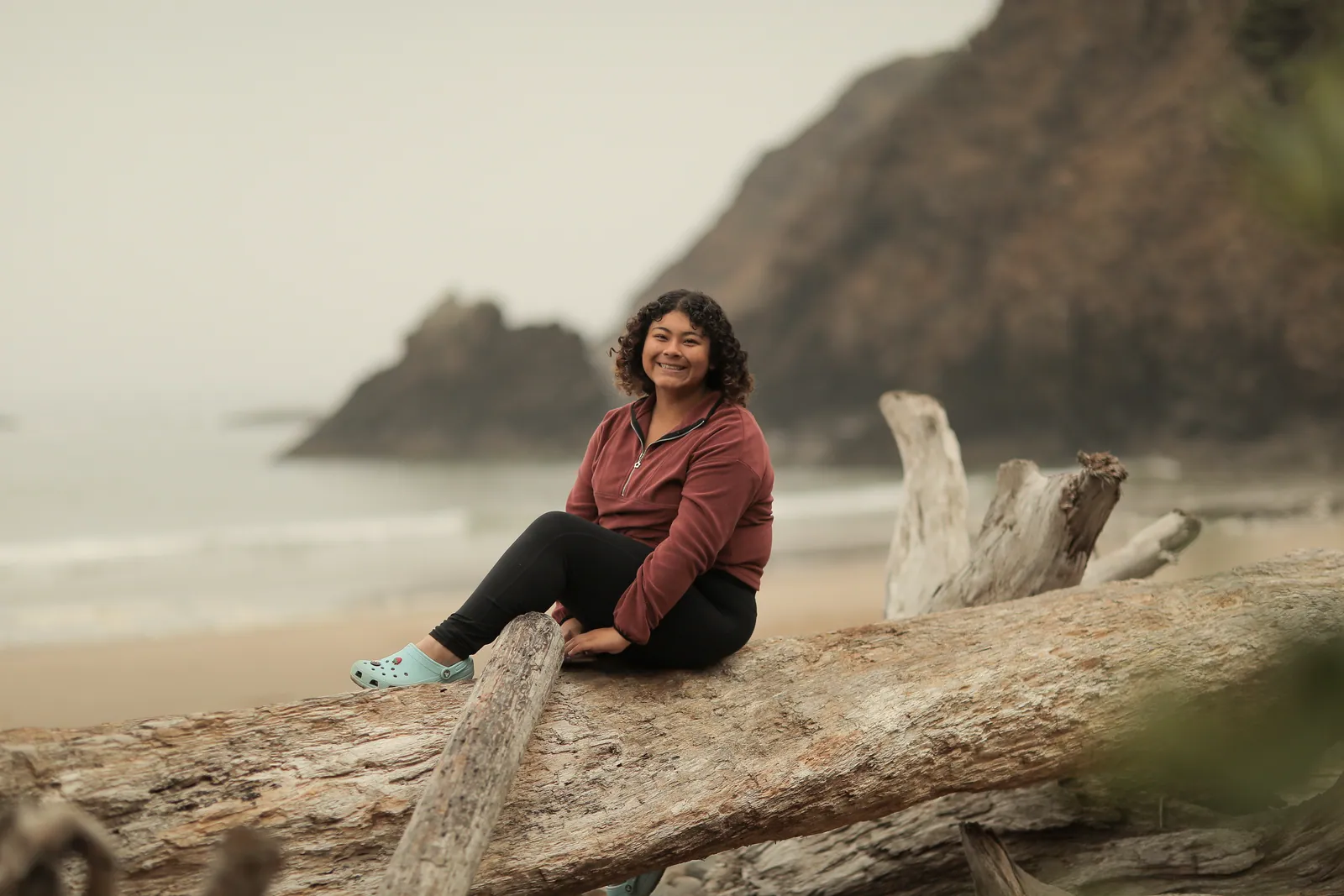 A smiling woman in a maroon fleece and mint clogs sits on a weathered driftwood log on a misty beach, with a sea stack and headland behind.