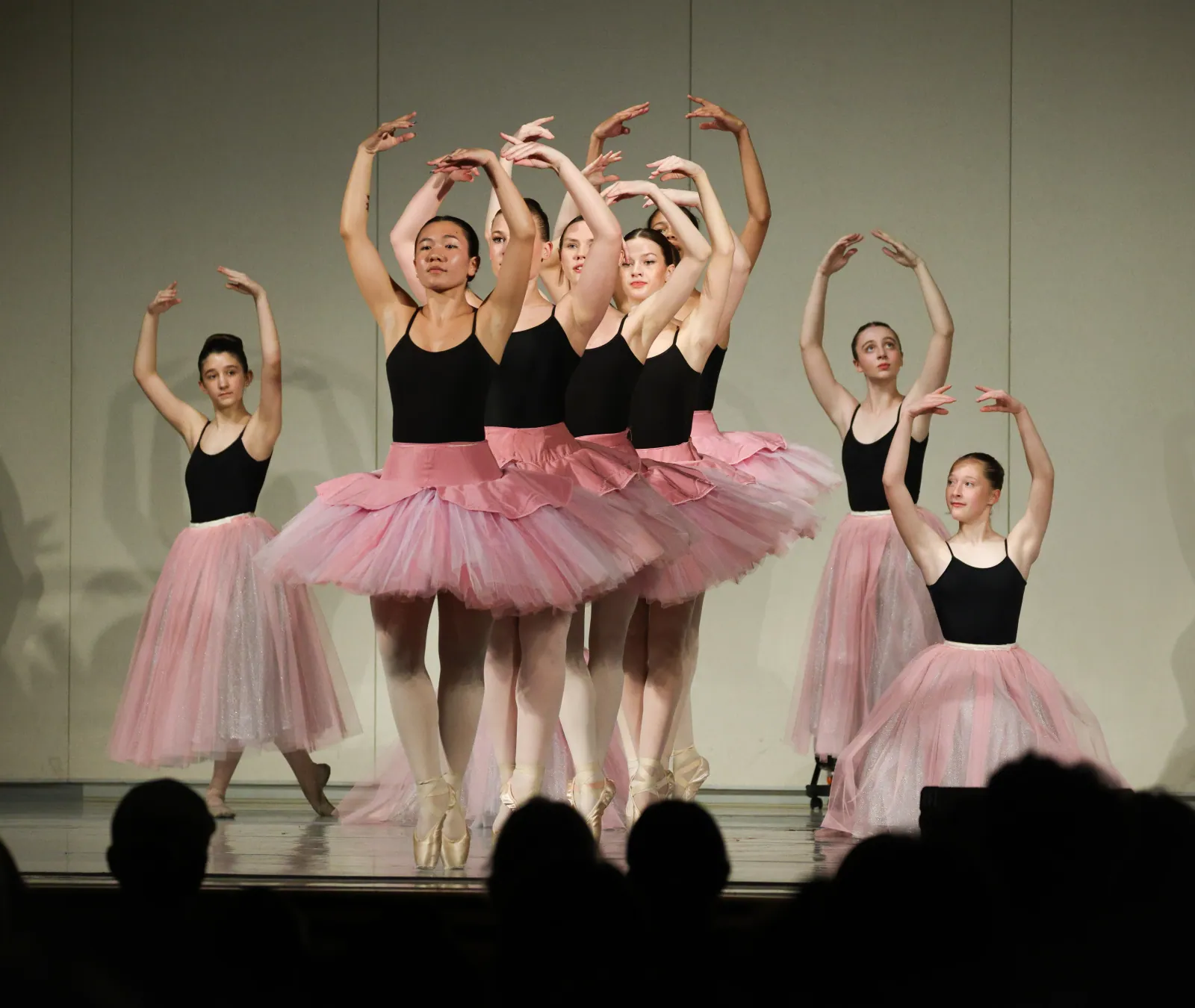 Young ballet dancers in black leotards and long pink tulle skirts hold arched arms overhead on pointe, framed by audience silhouettes.