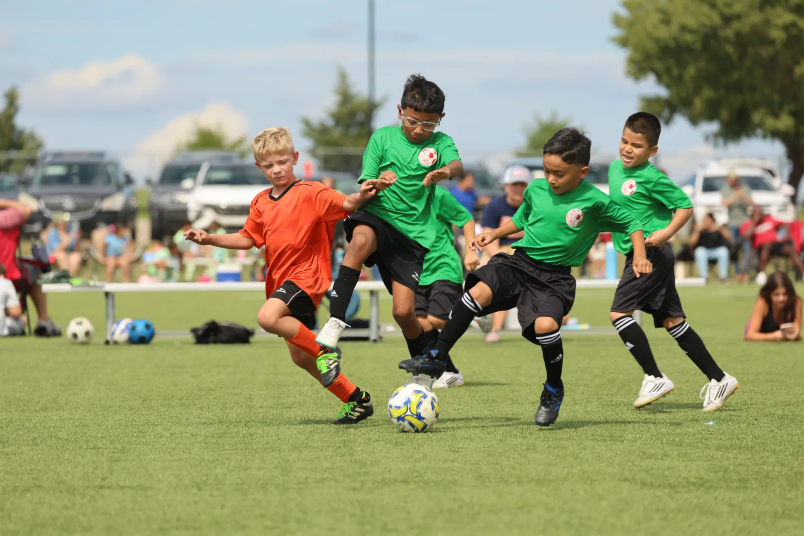 A boy in an orange kit battles three boys in green jerseys for a soccer ball on a sunny field, with parents watching from the sideline.