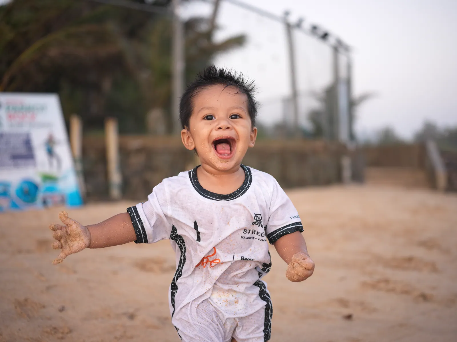 A toddler in a white jersey runs across the sand laughing with mouth wide open, hands caked in sand, with a blurred fence and trees behind.