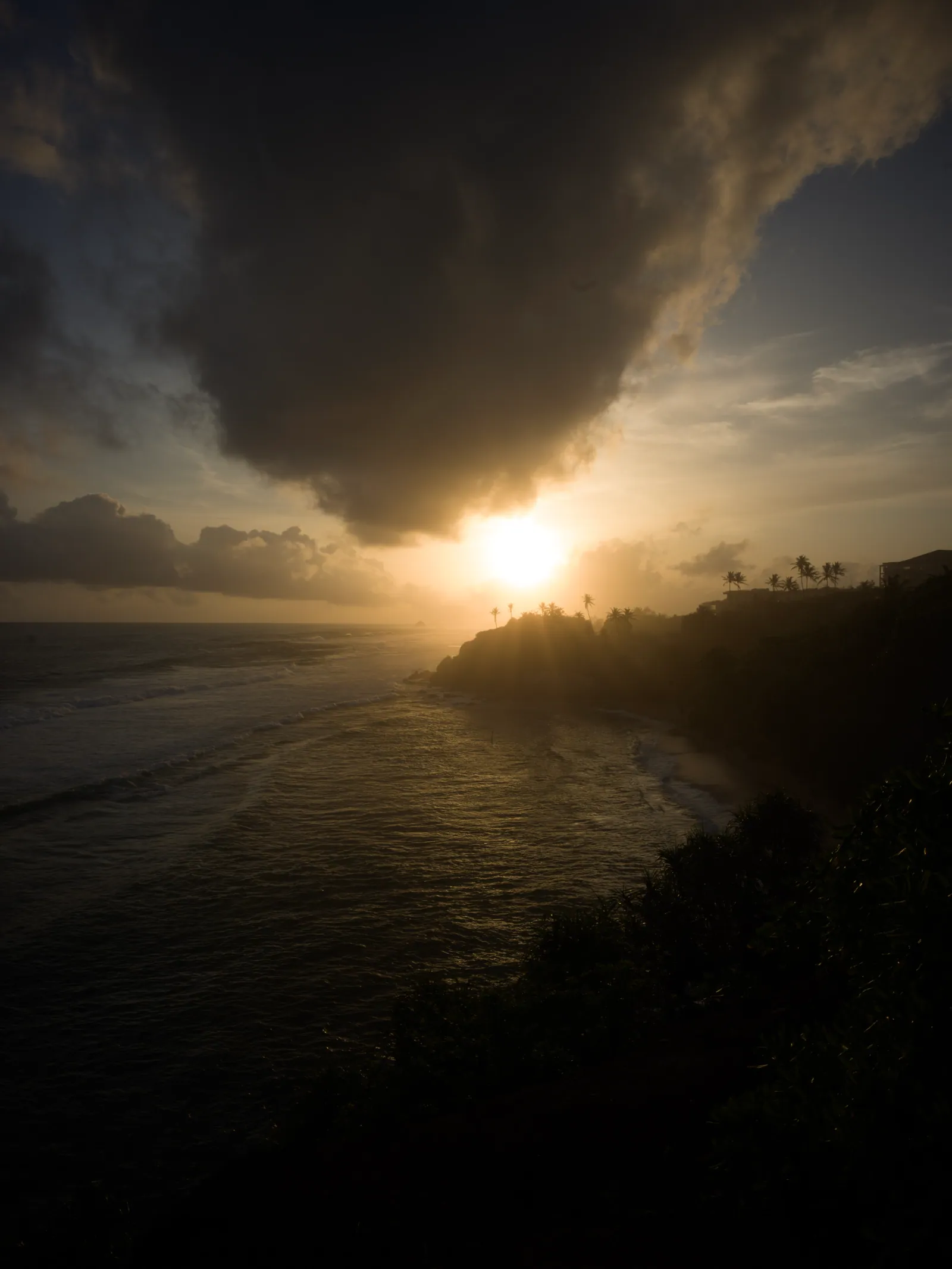 A heavy dark cloud hangs over a setting sun as golden light silhouettes a palm-topped headland and catches the waves of a quiet bay.
