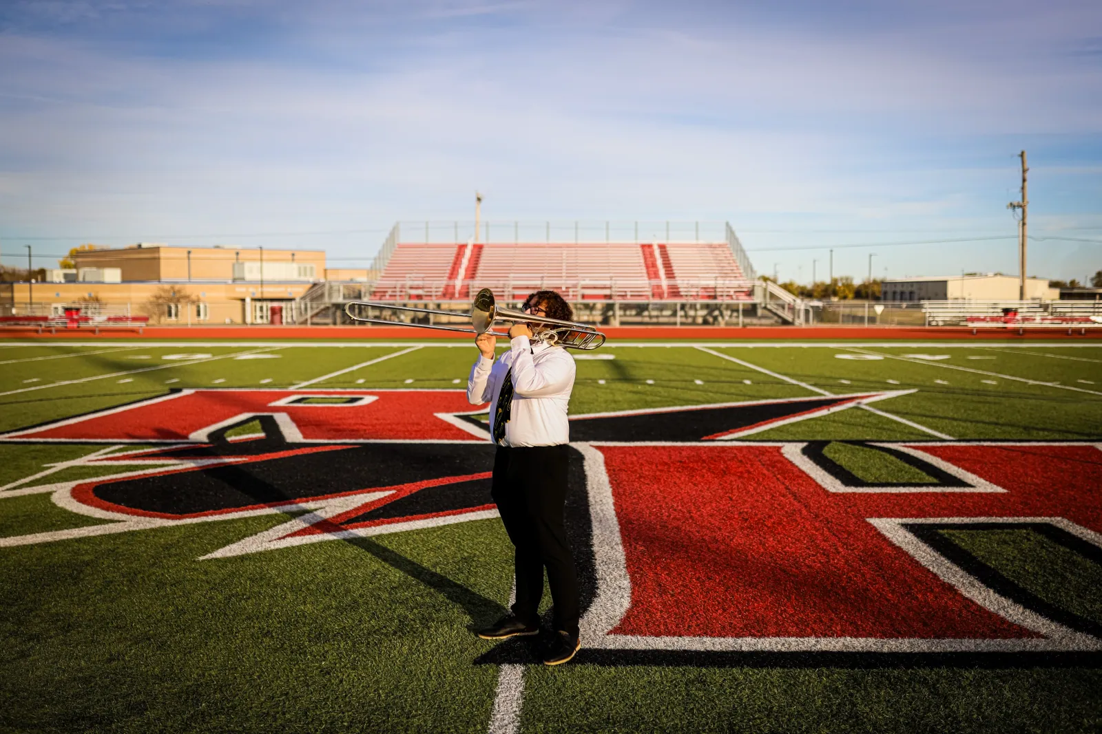 A young musician in a white shirt and tie plays a trombone on the red and black logo of a football field, with empty bleachers under blue sky.