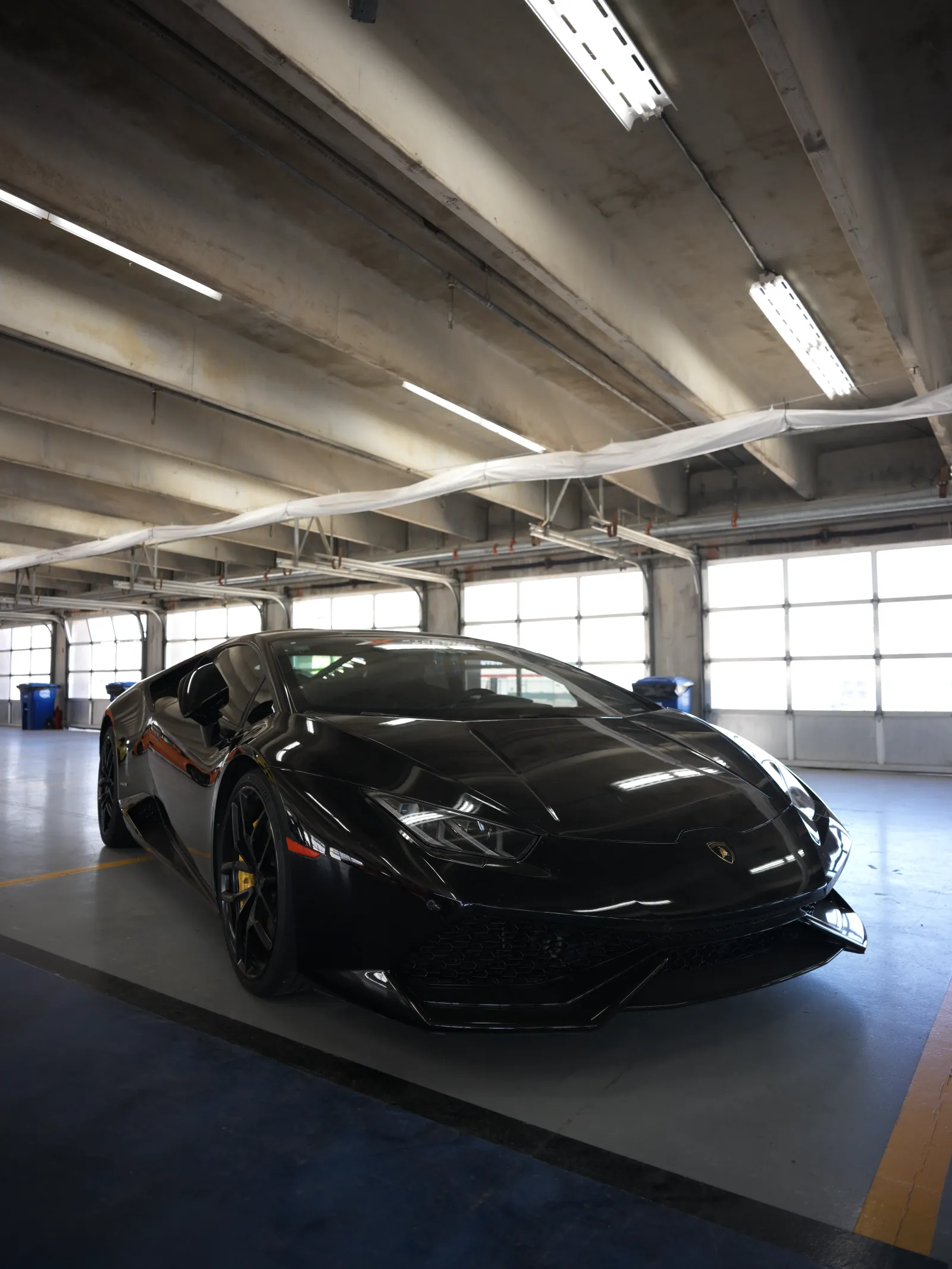 A glossy black Lamborghini sports car sits low in a concrete parking garage, lit by fluorescent tubes and bright windows behind it.