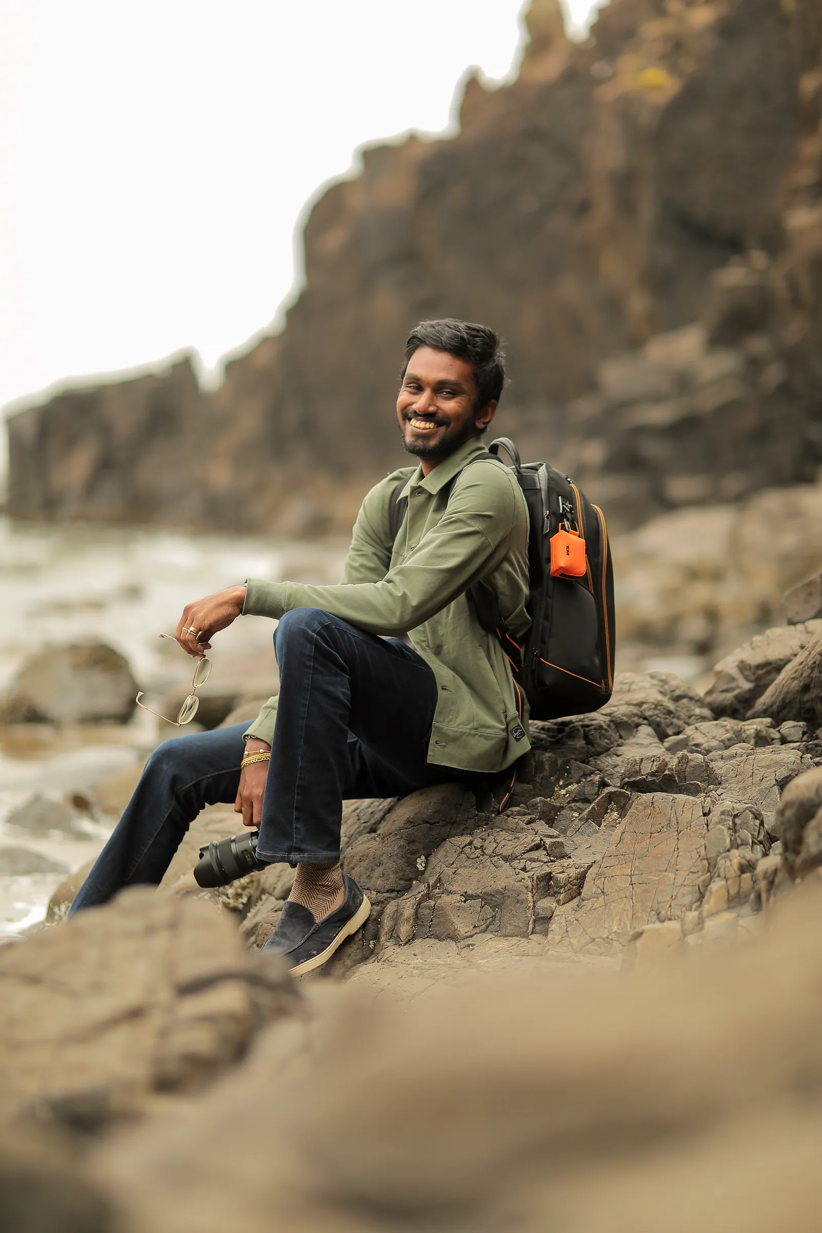 A smiling man in an olive shirt with a backpack sits on coastal rocks holding a camera and glasses, with a cliff and misty shore behind.