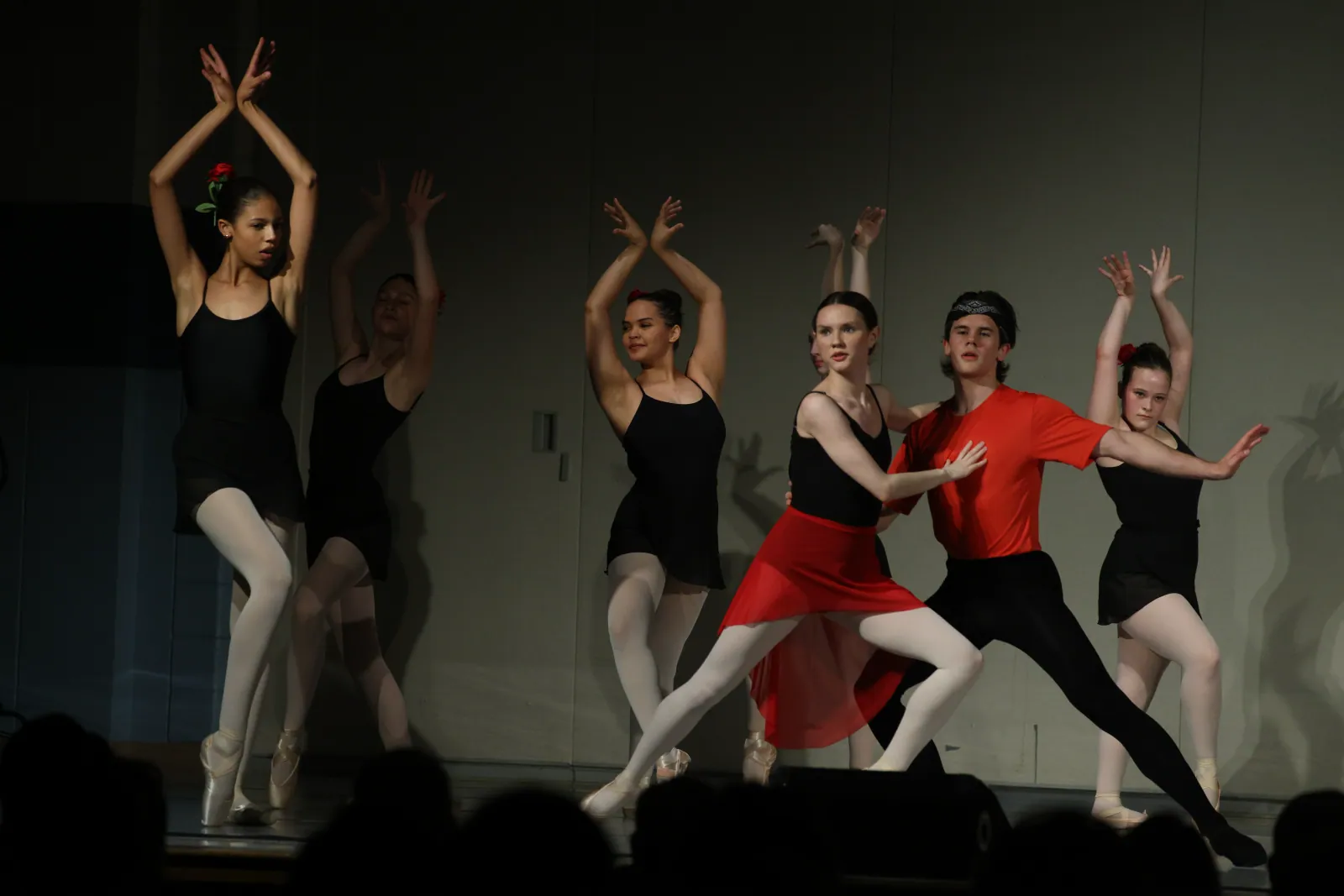 Ballet dancers in black leotards and white tights pose with arms crossed overhead while a pair in red lunges forward on a dimly lit stage.