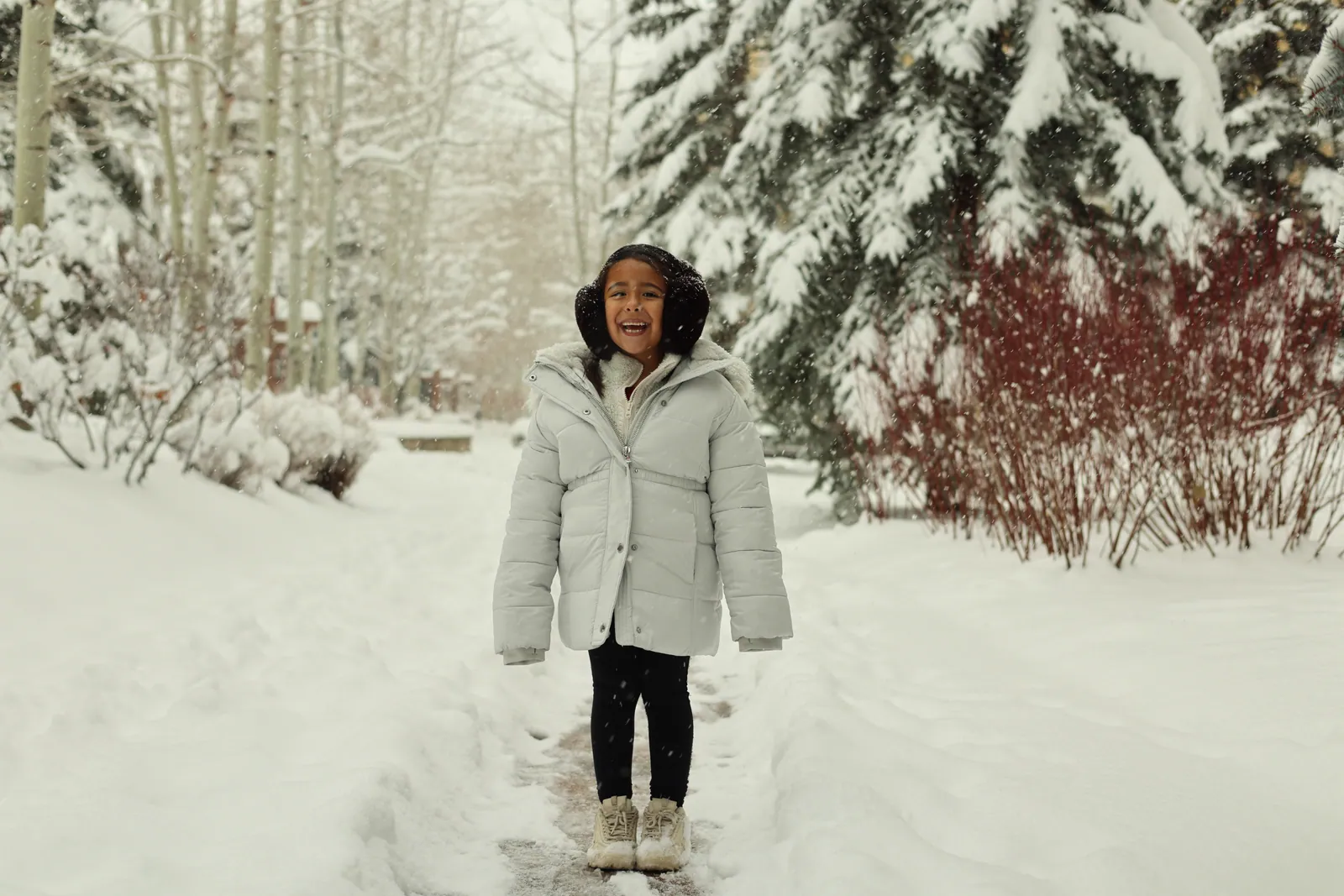 A laughing child in a pale puffer coat and earmuffs stands on a snowy path between snow-laden evergreens, aspens and red-stemmed shrubs.