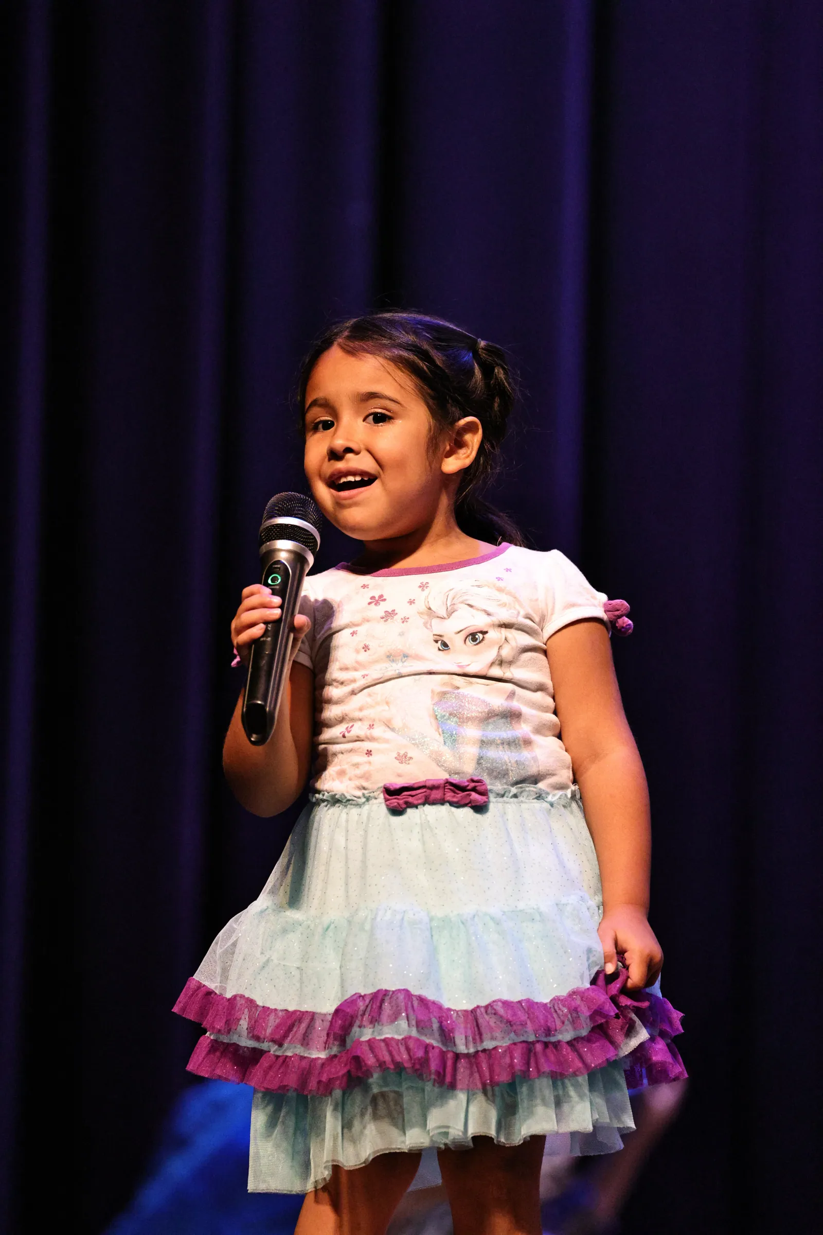 A young girl in a white top and a blue tulle skirt with purple ruffles sings into a microphone on stage in front of a deep blue curtain.