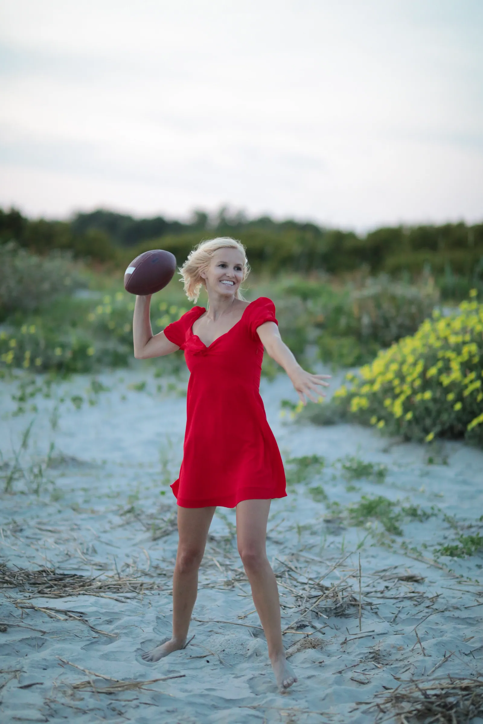 A smiling blonde woman in a red dress throws an American football barefoot on sandy dunes dotted with yellow wildflowers under a soft sky.