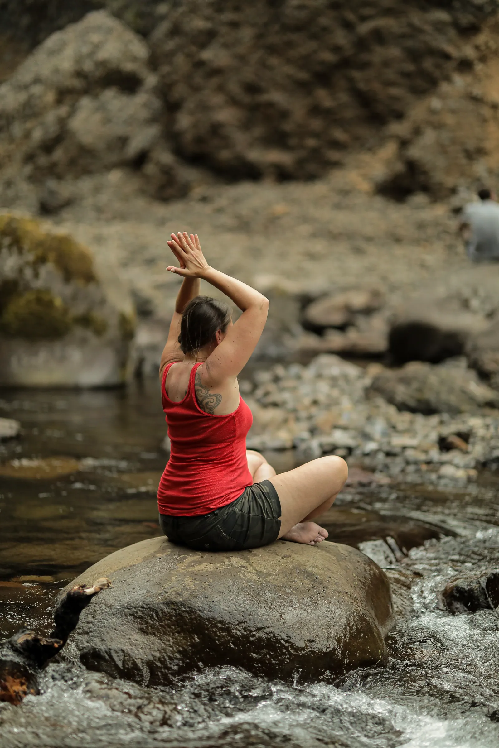 A woman in a red tank top sits cross-legged on a boulder in a rushing stream, arms raised overhead with palms together, facing the rocky bank.