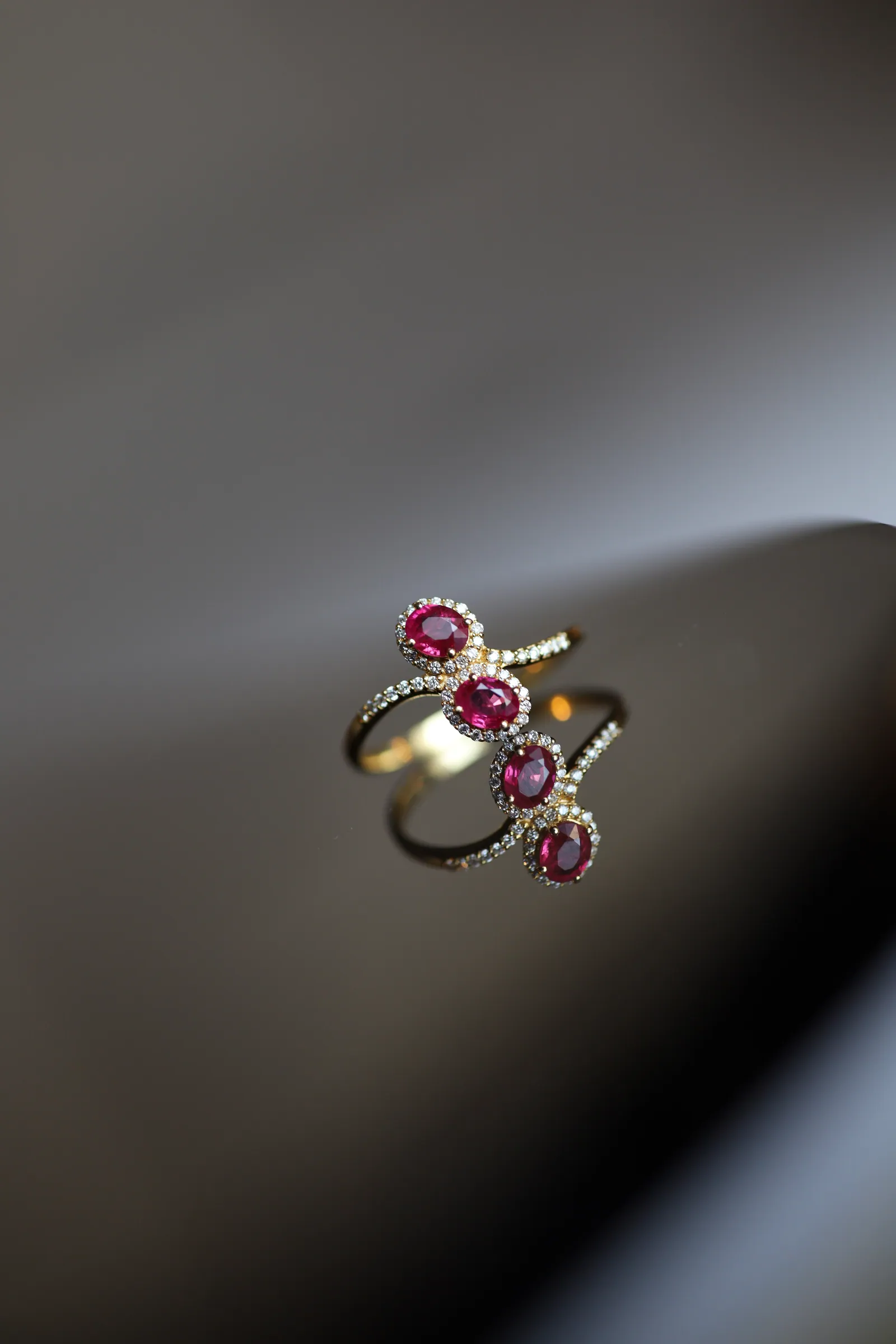 A gold ring set with four oval deep-pink gemstones, each ringed in tiny diamonds, resting on a glossy dark surface against soft grey.