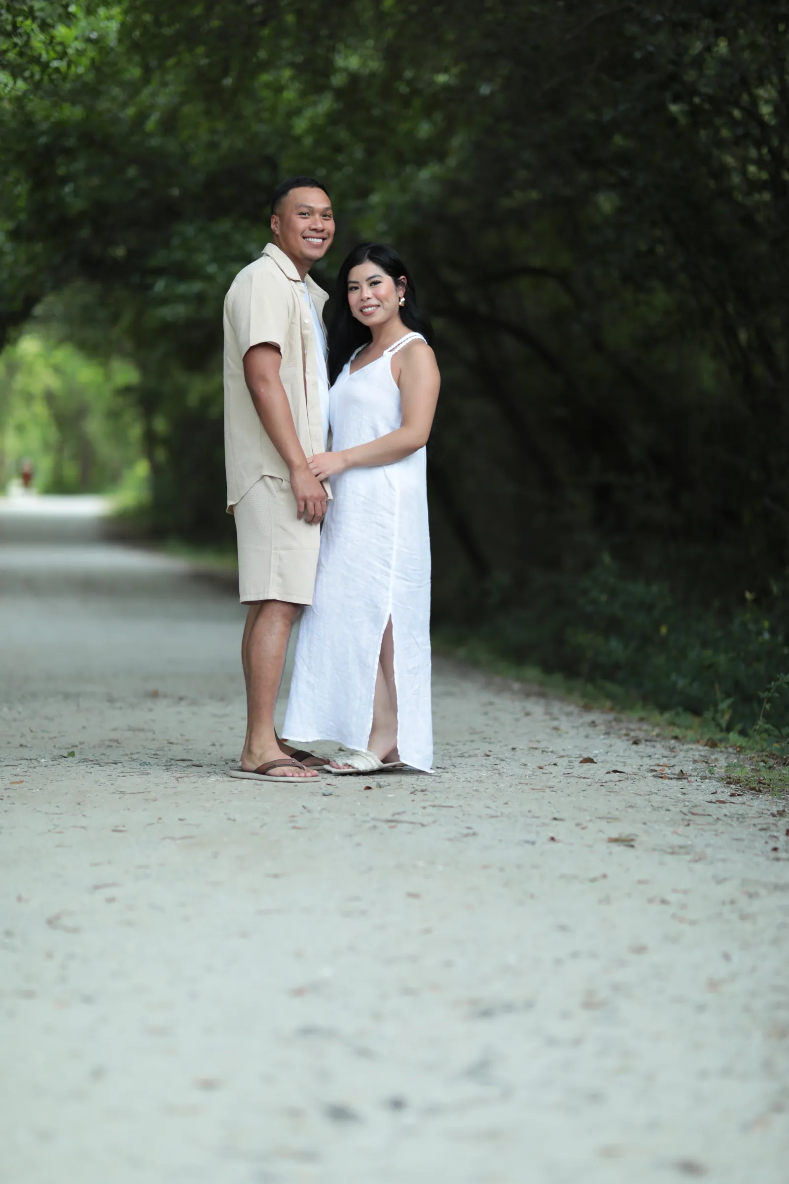 A smiling couple, he in beige and she in a long white dress, stand close together on a gravel path beneath an arching tunnel of trees.