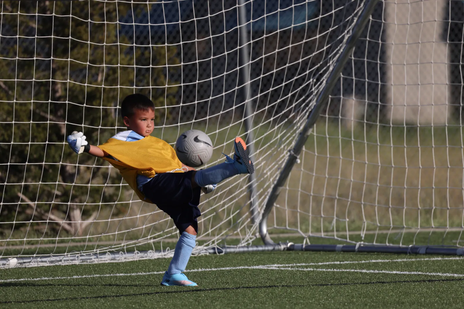 A young goalkeeper in a yellow bib and gloves swings his leg high to kick a soccer ball away from the net on a sunny artificial turf field.