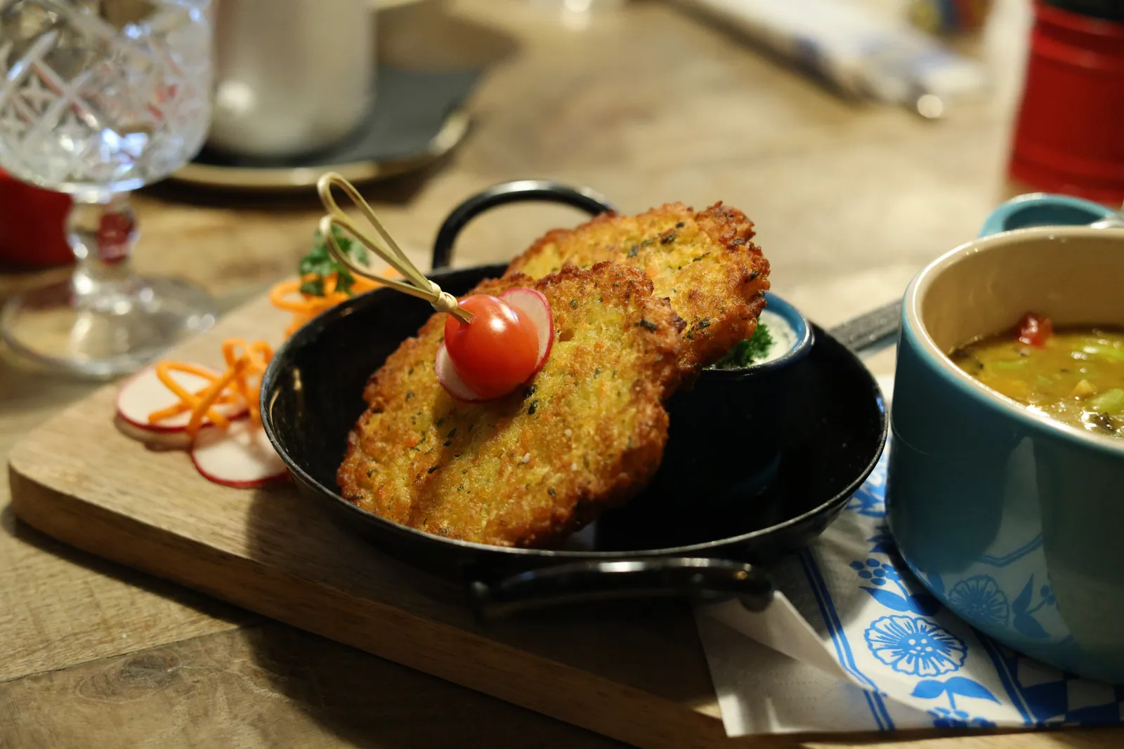Two crisp golden fritters in a small black skillet with dipping sauce and a tomato-radish pick, beside a blue bowl of stew on a wooden board.