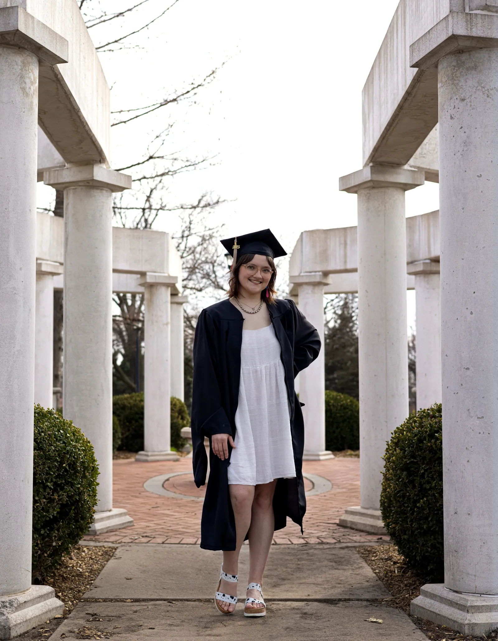 A smiling graduate in a black cap and gown over a white dress stands on a path between rows of white stone columns and rounded shrubs.