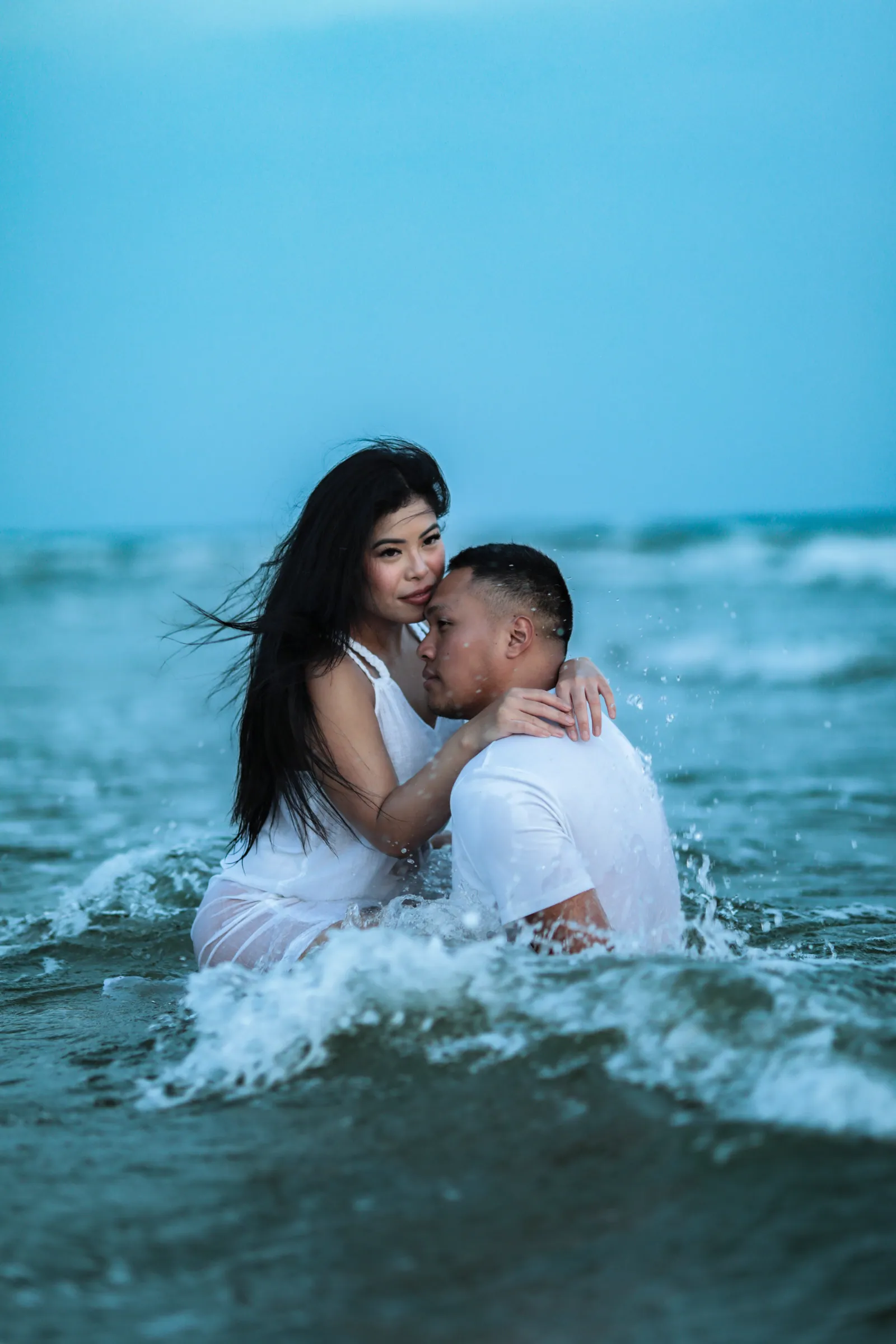 A couple dressed in white embrace waist-deep in the ocean at blue hour, her long dark hair blowing as a wave splashes around them.