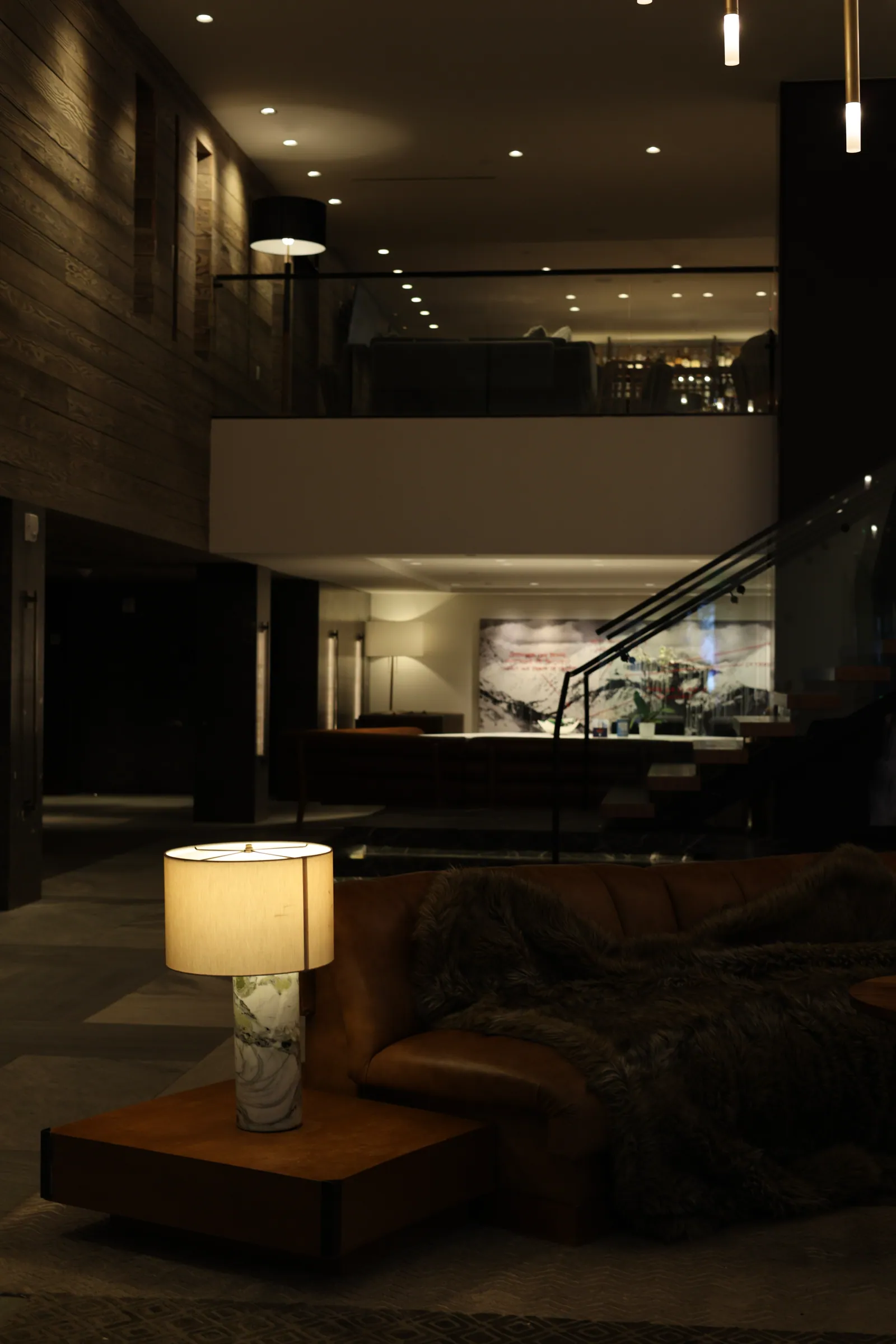 A dim lounge lit by a single table lamp beside a leather sofa draped in a fur throw, with a glass-railed mezzanine and stairs behind.
