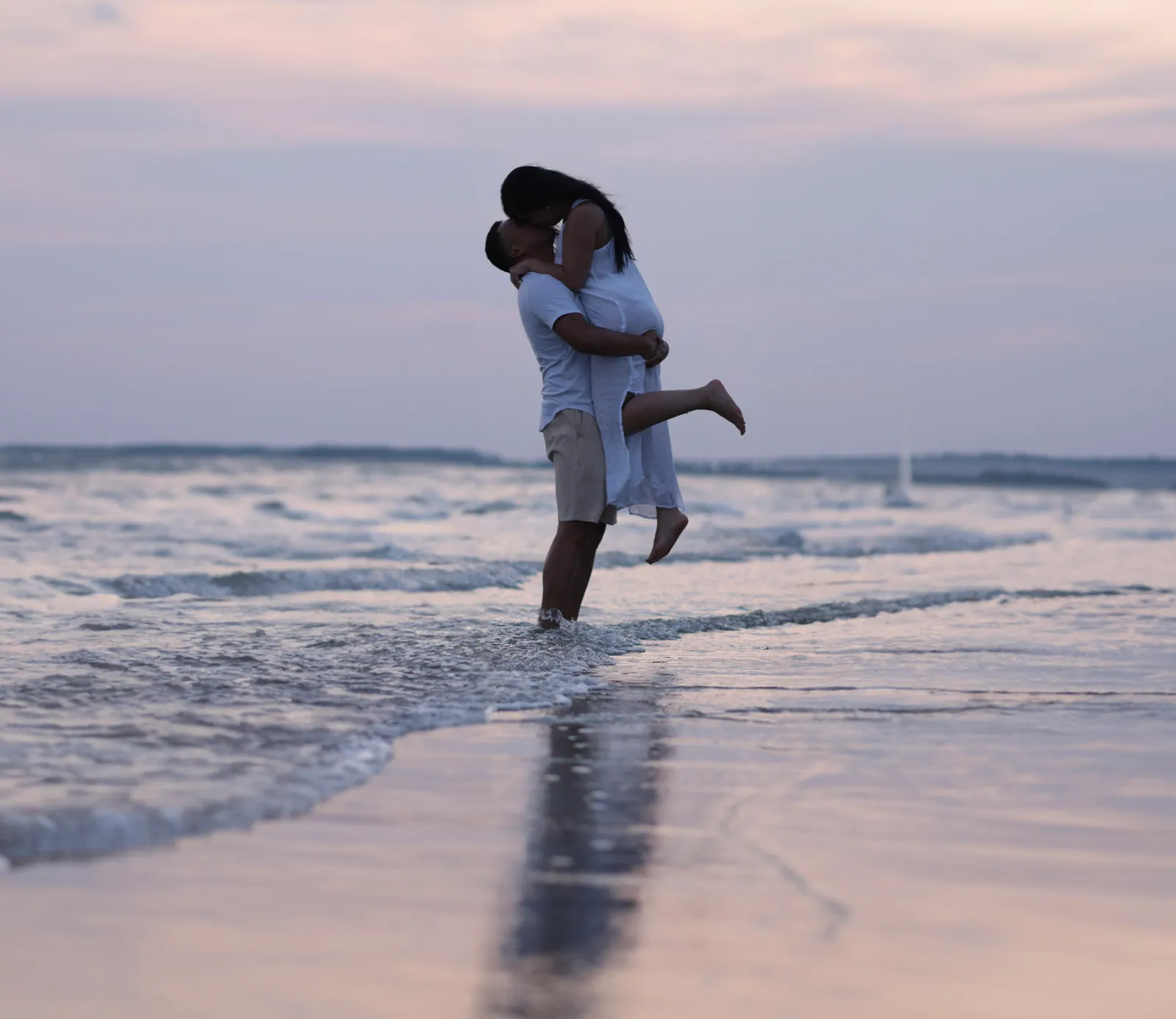 A man lifts a barefoot woman in a white dress for a kiss at the edge of the surf, their reflection on wet sand under a pale pink dusk sky.