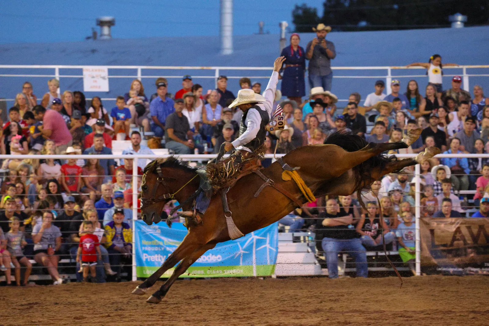 A rider in a white cowboy hat holds one arm high on a bucking brown horse in a dirt arena, packed stands of spectators behind him at dusk.