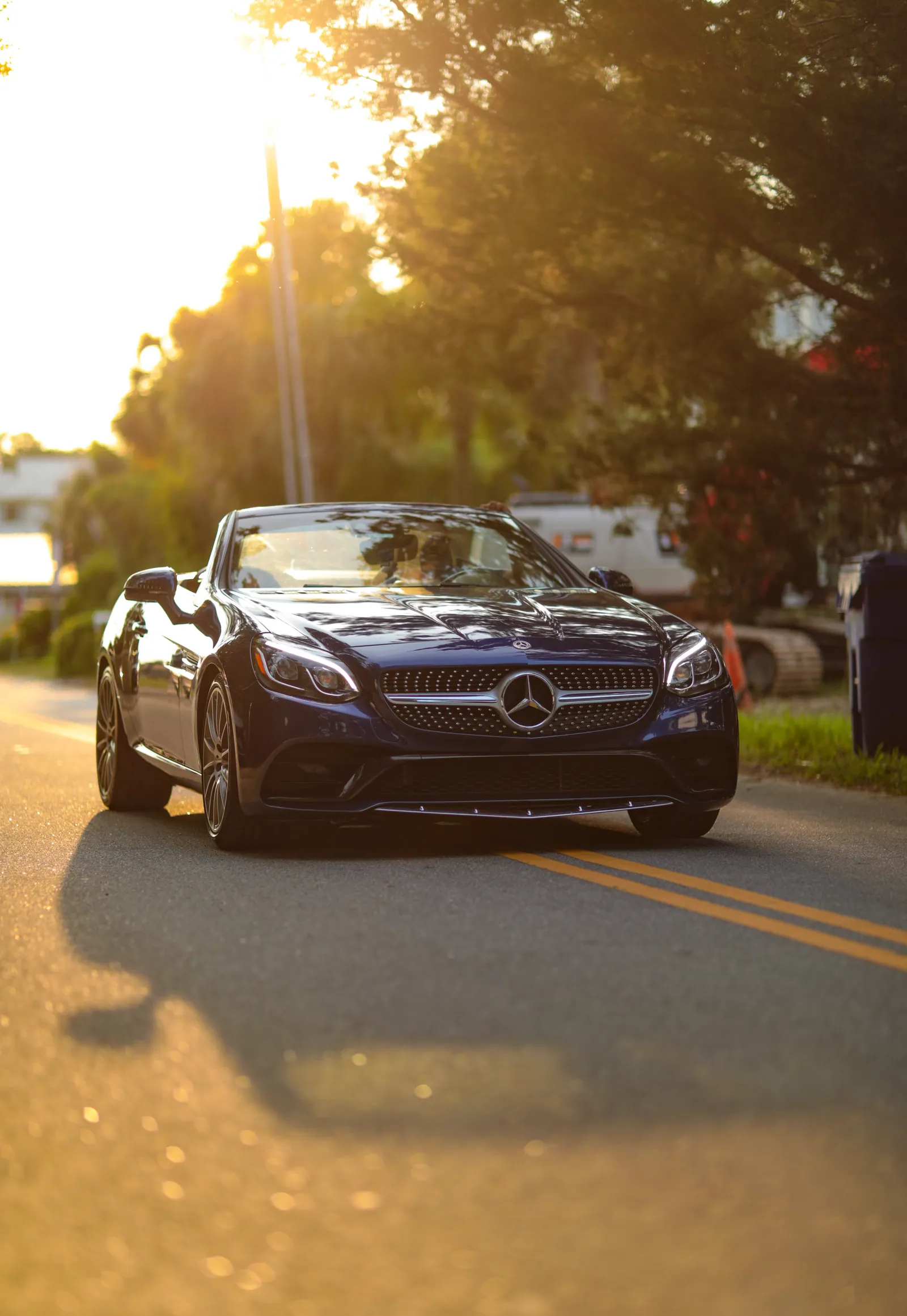 A dark blue Mercedes convertible rolls along a quiet road at golden hour, sunlight flaring through trees and its shadow stretching across the asphalt.