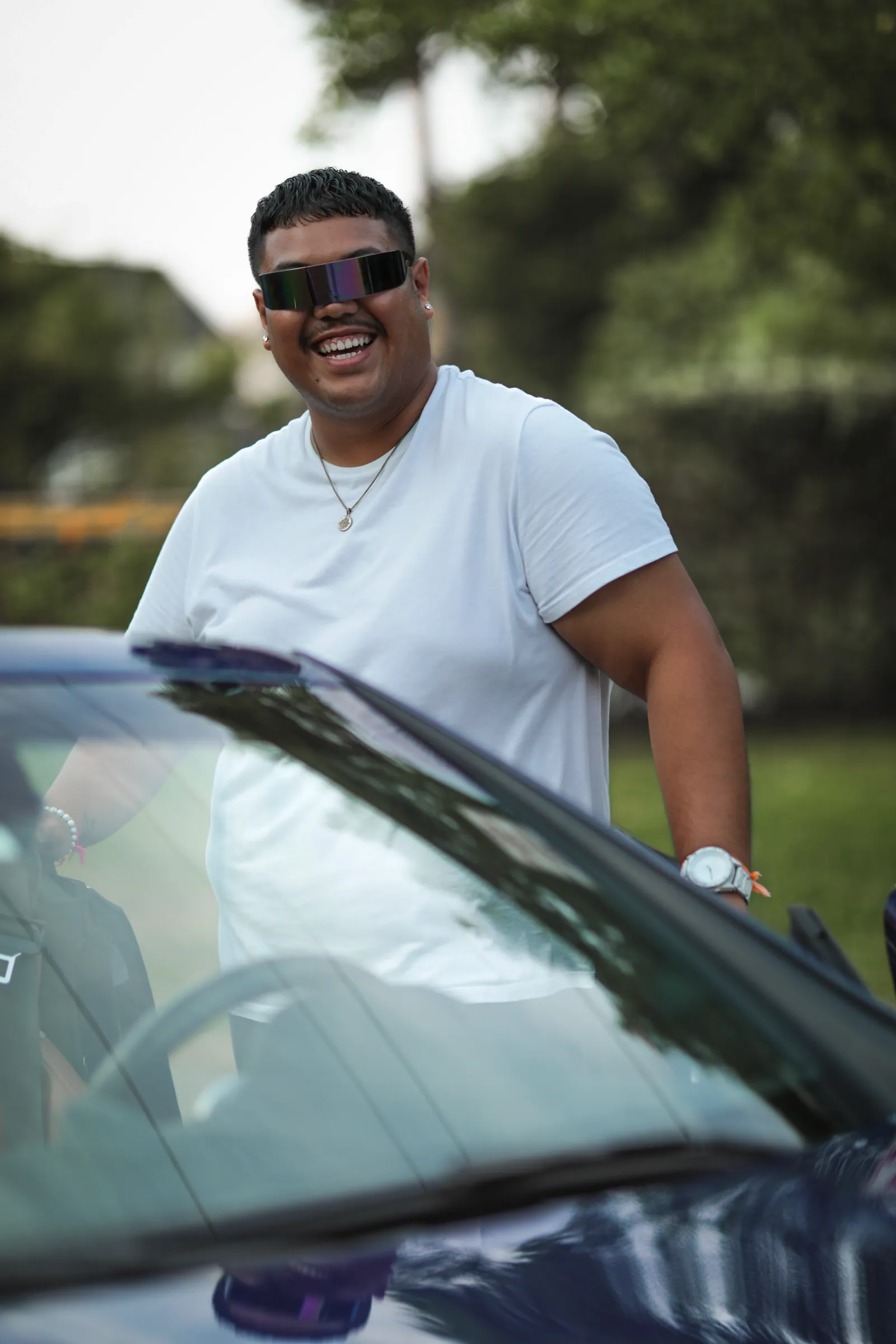 A laughing man in a white T-shirt and reflective visor sunglasses leans over the door of a dark open-top car, with soft green trees behind.