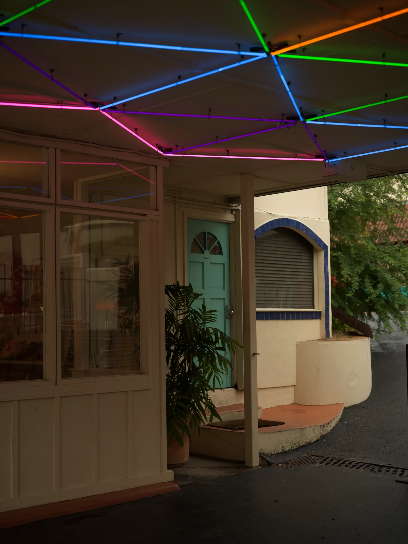 Pink, blue, green and orange neon tubes form a geometric web under a canopy above a cream building with a teal door and tiled arched window.