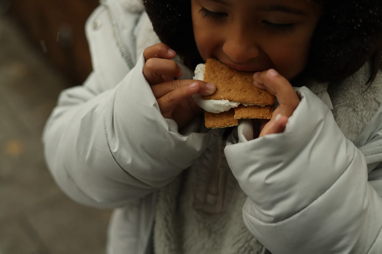 A child in a pale puffer coat, eyes closed, takes a big bite of a s'more made with graham crackers and marshmallow as light snow falls.
